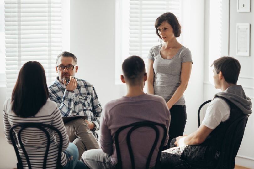 Five adults sit and stand in a circle of black chairs while one man holds a pen near his face and a clipboard in hand.