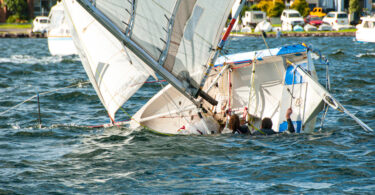 Two people are in a body of water holding onto their capsized sailboat. There are other boats in distance.