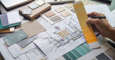 A close-up of an interior designer looking at paint swatches and room sketches laid out on a desk near a laptop.