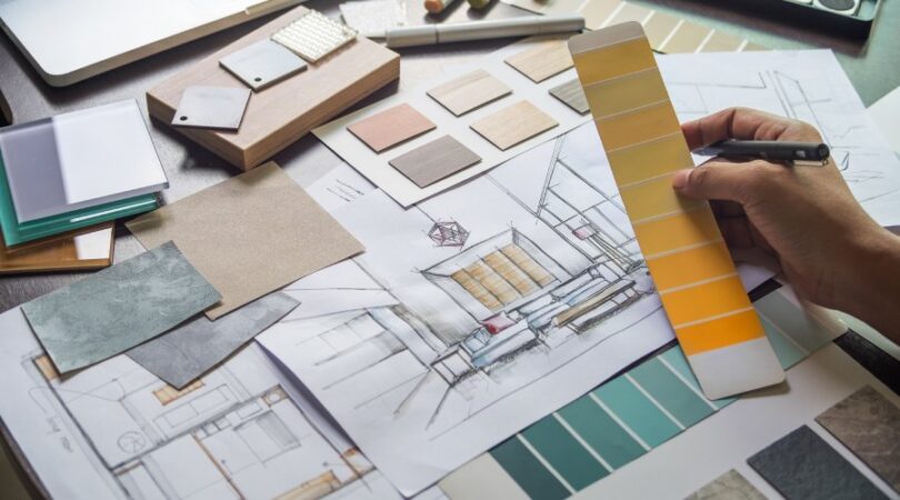 A close-up of an interior designer looking at paint swatches and room sketches laid out on a desk near a laptop.