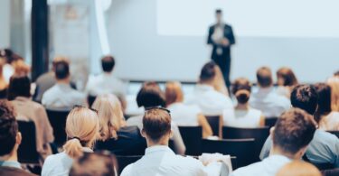 An audience sits in black chairs as they look and listen to a speaker standing on a stage for an event.