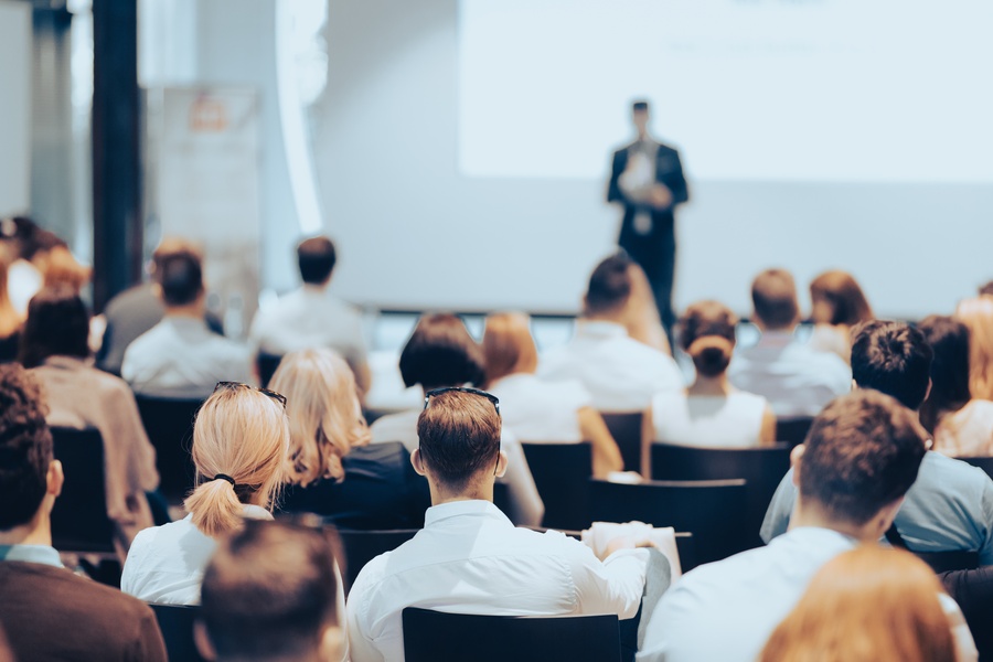 An audience sits in black chairs as they look and listen to a speaker standing on a stage for an event.
