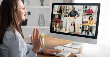 A woman sits at a computer desk and waves to a screen of remote workers. Some of the workers wave back to her.