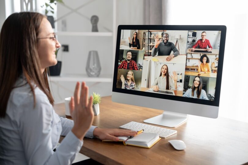 A woman sits at a computer desk and waves to a screen of remote workers. Some of the workers wave back to her.
