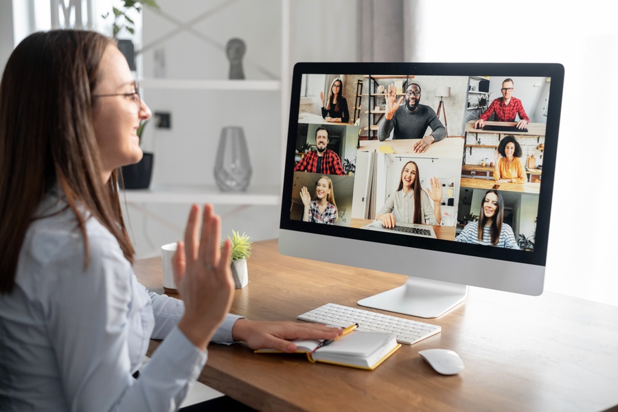 A woman sits at a computer desk and waves to a screen of remote workers. Some of the workers wave back to her.