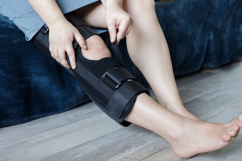 A close-up view shows a person sitting on a blue couch, adjusting the velcro on a black brace around their knee.