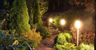 Three lights shine bright in a home's garden around a brick pathway. Several trees stand around the lights as well.
