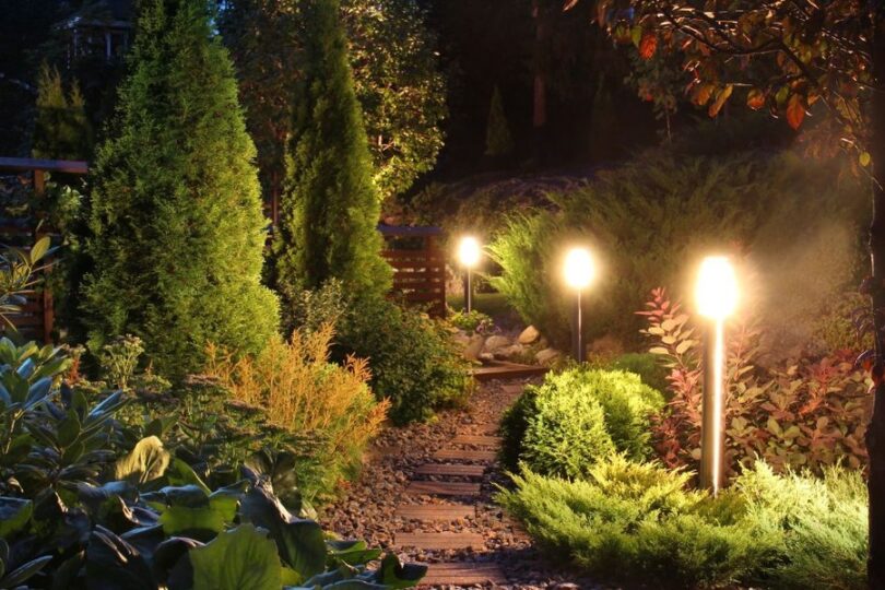 Three lights shine bright in a home's garden around a brick pathway. Several trees stand around the lights as well.