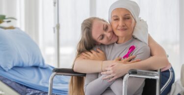 A middle-aged woman in a wheelchair as a friend hugs her from behind. The patient has a pink ribbon on her shirt.