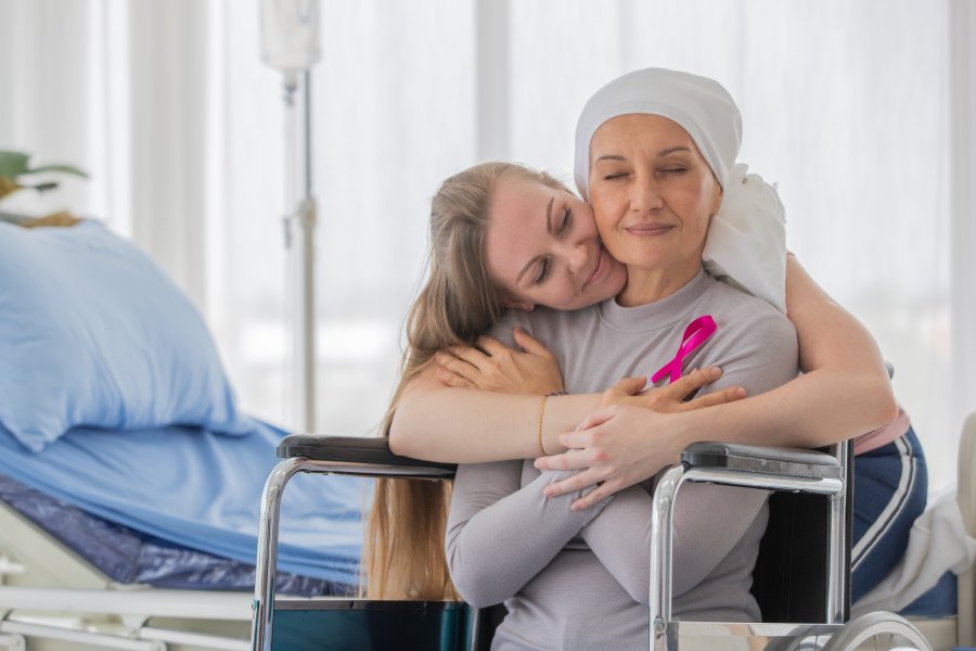 A middle-aged woman in a wheelchair as a friend hugs her from behind. The patient has a pink ribbon on her shirt.
