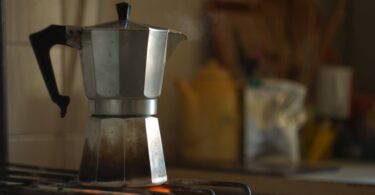 A stainless steel Moka pot placed on the stovetop burner. The flame is burning underneath and the pot has a black handle.