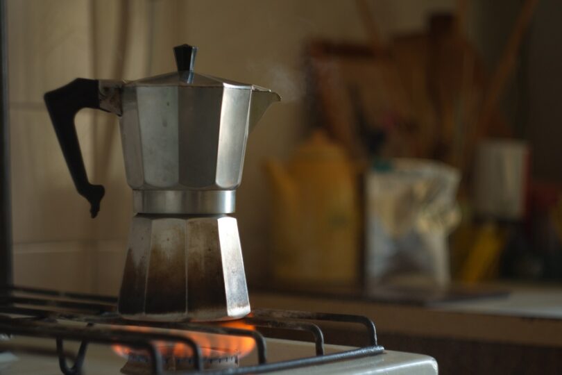 A stainless steel Moka pot placed on the stovetop burner. The flame is burning underneath and the pot has a black handle.