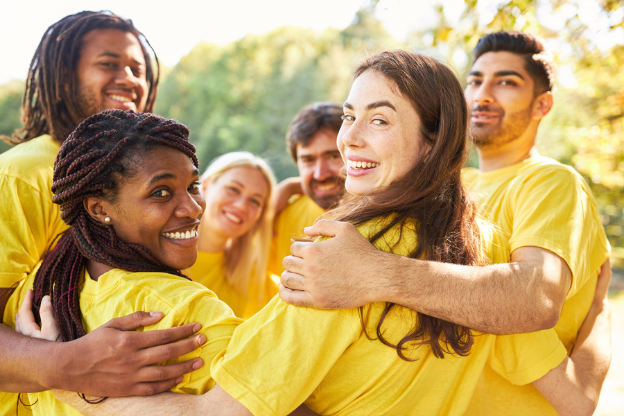 A group of smiling men and women in yellow shirts standing in a circle outdoors and hugging each other.