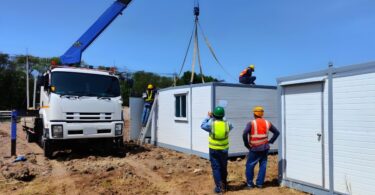 A group of workers in safety vests and hard hats using a tall crane to position a white shipping container.