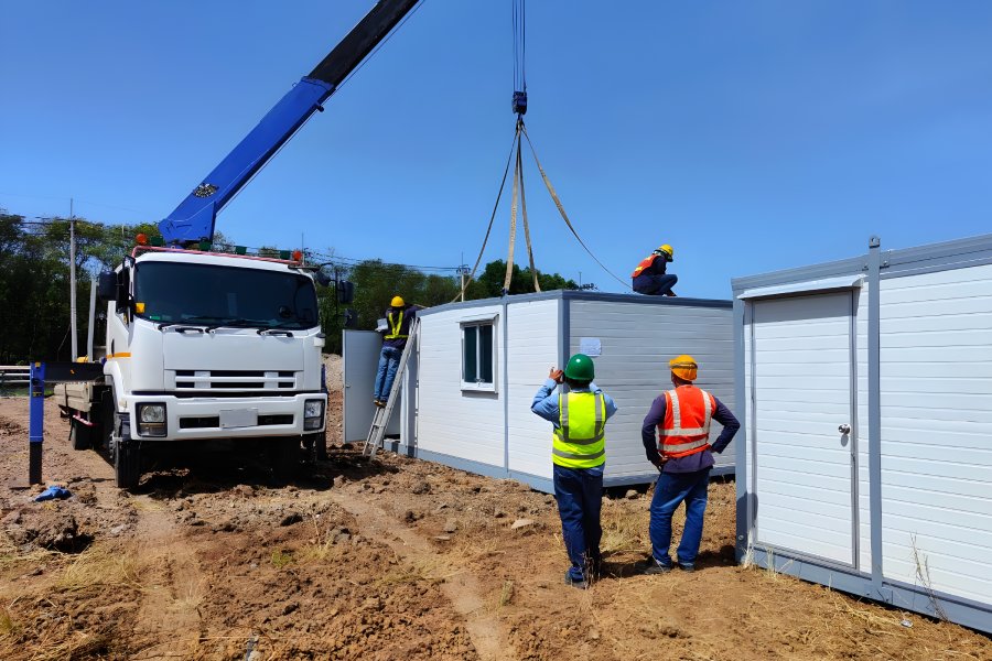 A group of workers in safety vests and hard hats using a tall crane to position a white shipping container.