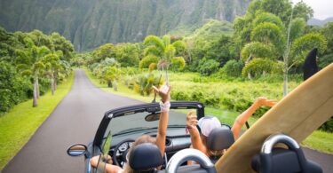 Two girls are driving down a road in Hawaii with their hands up, the car's roof down, and a surfboard in the back seat.