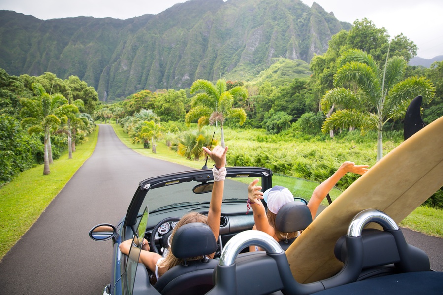 Two girls are driving down a road in Hawaii with their hands up, the car's roof down, and a surfboard in the back seat.