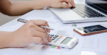 A close-up of someone's hand using a calculator and a laptop, referencing a paper graph data while doing finance.