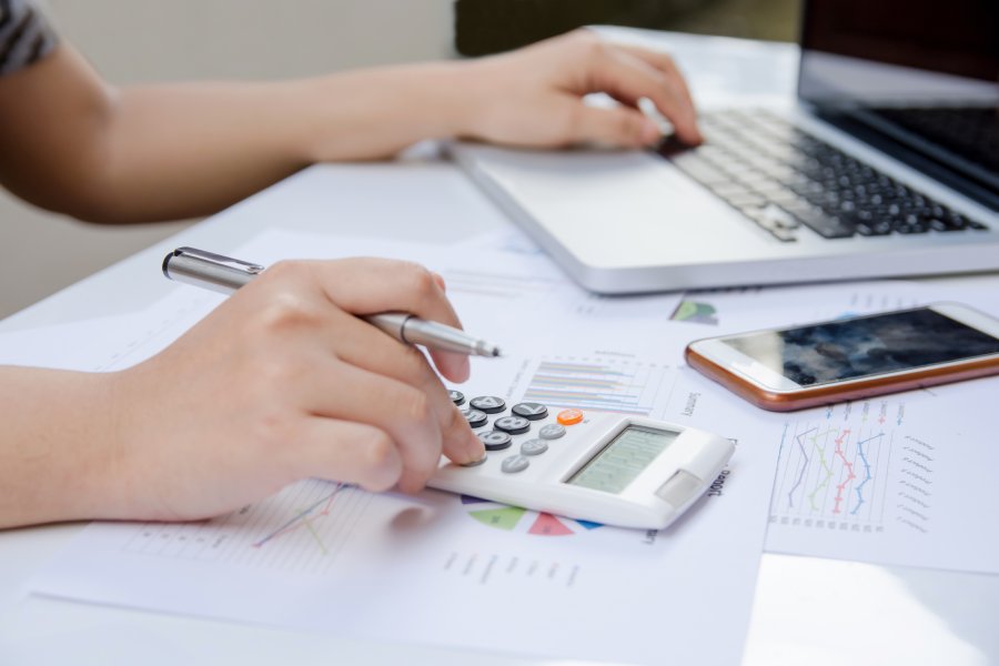A close-up of someone's hand using a calculator and a laptop, referencing a paper graph data while doing finance.