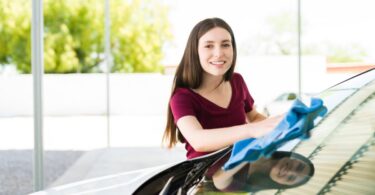 A woman smiles as she washes her car, using a blue microfiber cloth to wipe water off the front windows.
