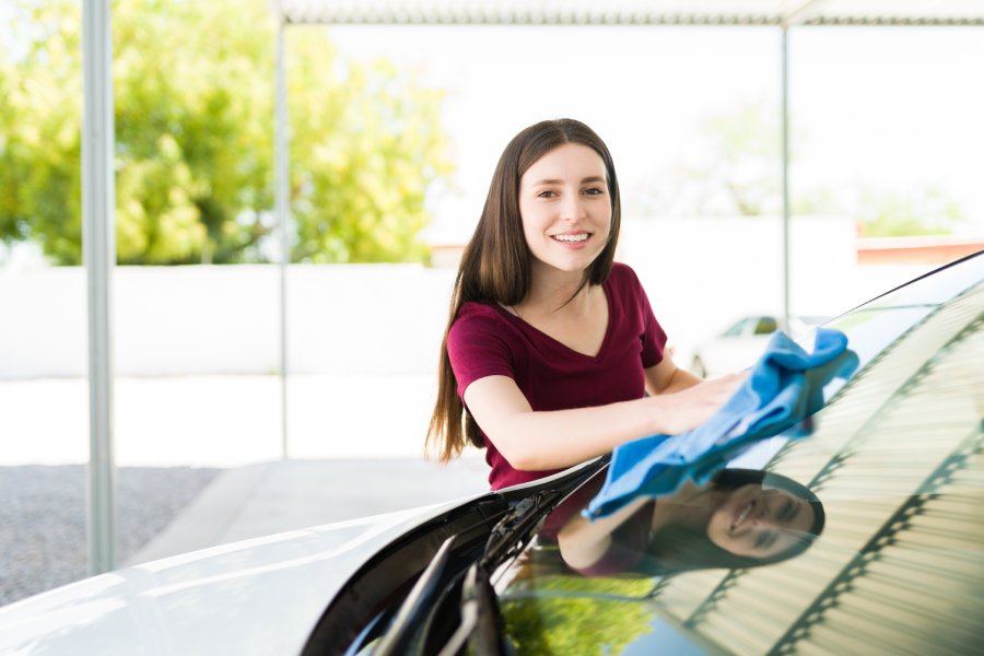 A woman smiles as she washes her car, using a blue microfiber cloth to wipe water off the front windows.
