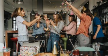 A group of teenagers gathered around one teen at a party. They are tossing paper confetti into the air to celebrate.