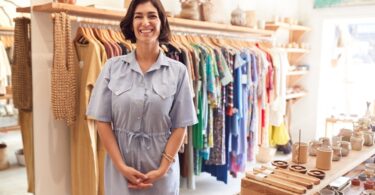 A smiling woman in a blue dress is standing in a small boutique. Behind her is a rack of clothing items.