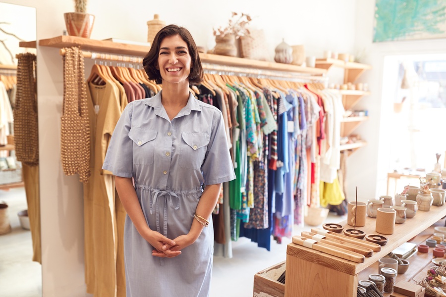 A smiling woman in a blue dress is standing in a small boutique. Behind her is a rack of clothing items.