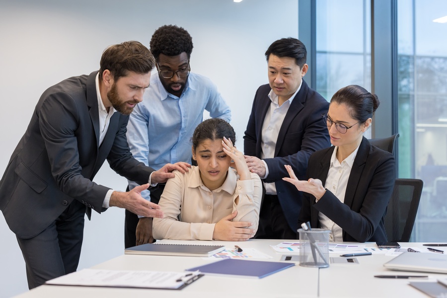 In an office space, a woman sits at a table and is overwhelmed, surrounded by other works talking to her.