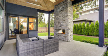 A luxury home features a spacious covered patio with a built-in outdoor fireplace. A row of bushes lines the fence.