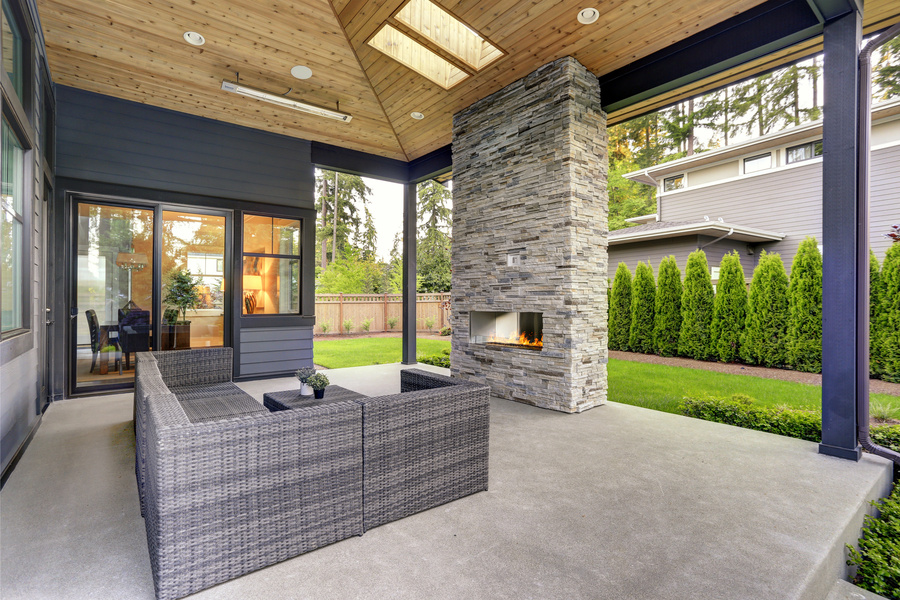 A luxury home features a spacious covered patio with a built-in outdoor fireplace. A row of bushes lines the fence.