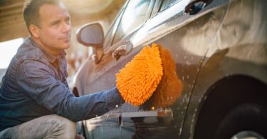 A man washing his car using a bright orange washing mitt. The car has soap suds and water streaks around the exterior.