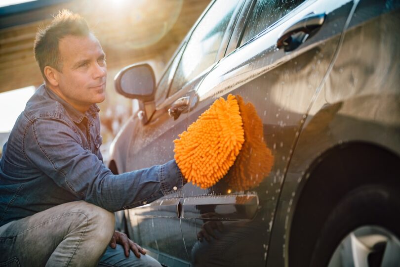 A man washing his car using a bright orange washing mitt. The car has soap suds and water streaks around the exterior.