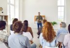 A person leading a corporate staff training in front of several rows of employees in workplace casual attire.