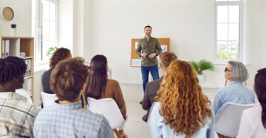 A person leading a corporate staff training in front of several rows of employees in workplace casual attire.