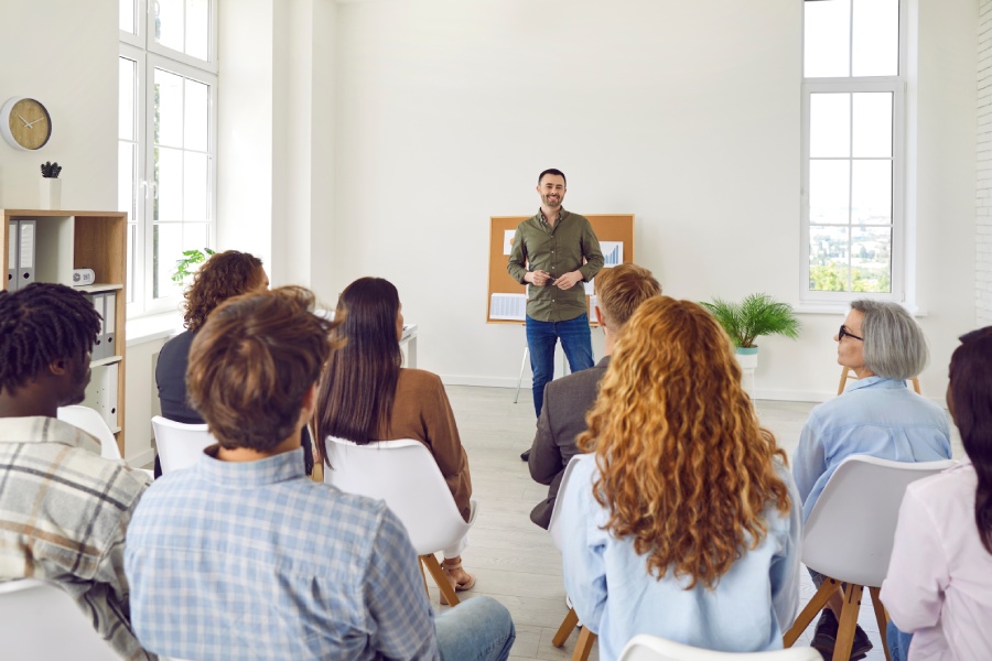 A person leading a corporate staff training in front of several rows of employees in workplace casual attire.