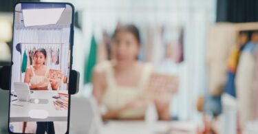 A social media influencer recording a product review while sitting at a white desk while a smartphone records the review.