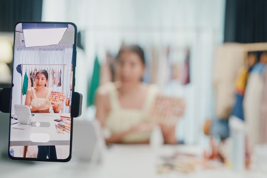 A social media influencer recording a product review while sitting at a white desk while a smartphone records the review.