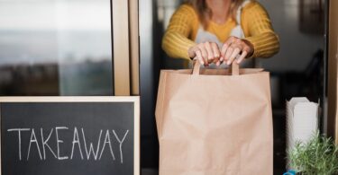 A blonde woman in a yellow sweater places a brown paper bag on a windowsill next to a chalkboard labeled “Takeaway.”
