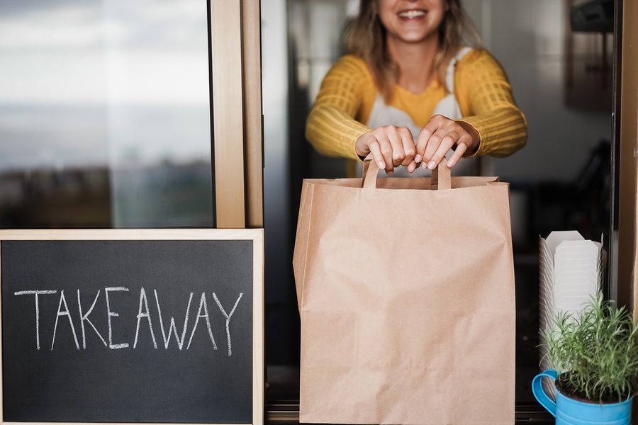 A blonde woman in a yellow sweater places a brown paper bag on a windowsill next to a chalkboard labeled “Takeaway.”