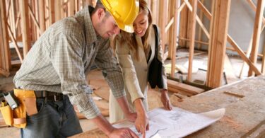 A man in a hard hat stands at a table with a young woman. They stand in an unfinished house, looking at blueprints.