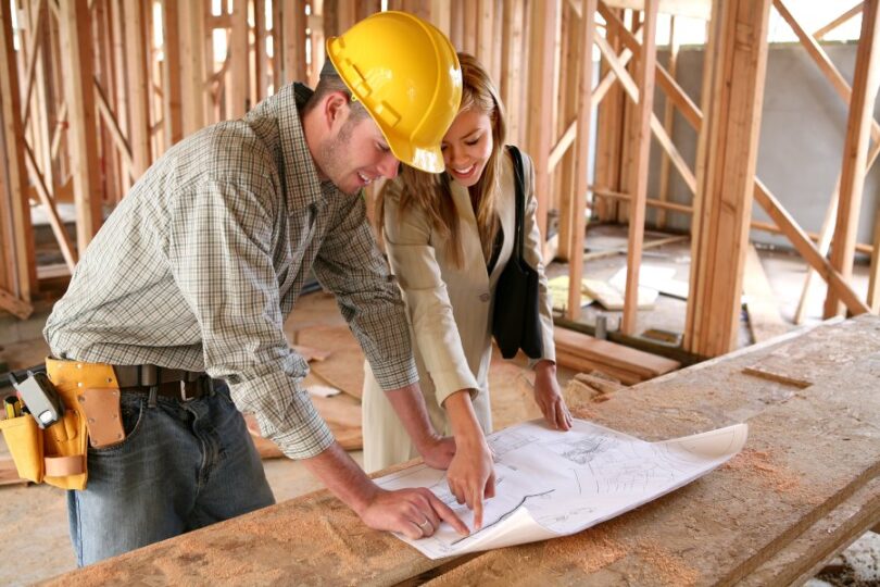 A man in a hard hat stands at a table with a young woman. They stand in an unfinished house, looking at blueprints.