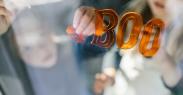 Two children decorate a window with orange stickers that spell the word "boo" and a pumpkin sticker.