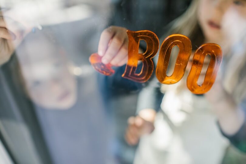 Two children decorate a window with orange stickers that spell the word "boo" and a pumpkin sticker.