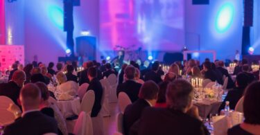 People sit around a bunch of tables in an event space. They all wear formal clothing and look toward the stage.