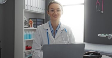 A smiling physician sits behind a laptop. Behind her sits a stock of chemical bottles and first aid items.