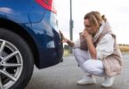 A confused woman looking at the bottom of the rear bumper of her car. She is crouched down next to the vehicle.