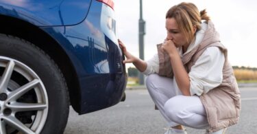 A confused woman looking at the bottom of the rear bumper of her car. She is crouched down next to the vehicle.