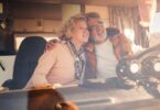A senior man grips the steering wheel of a camper van while embracing a senior woman in the passenger seat.
