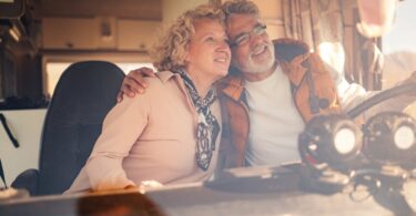 A senior man grips the steering wheel of a camper van while embracing a senior woman in the passenger seat.